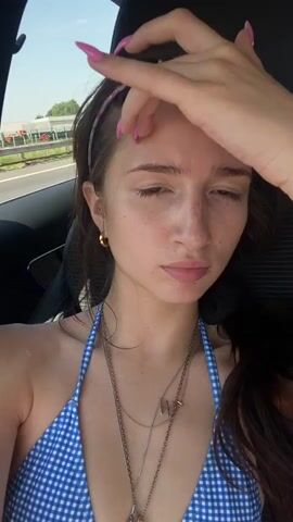The video shows a young woman with long, dark hair and a blue and white checkered bikini top, sitting in the passenger seat of a car. She is wearing a pink headband and several necklaces. The camera is angled downwards, showing her face from the chest up. Outside the car window, a road and trees are visible. She is looking at the camera and her hand is raised to her hair.