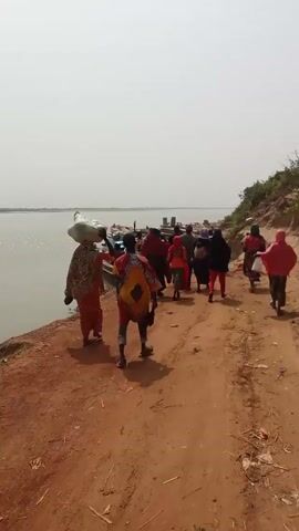 A group of people walks along a dirt path towards the edge of a wide body of water, possibly a river or lake, where several wooden boats are docked. The people appear to be of African descent, wearing a mix of traditional and modern clothing. One person carries a large sack on their head. The path is reddish-brown dirt and the water is grey with some white birds visible. The sky is overcast and light grey.