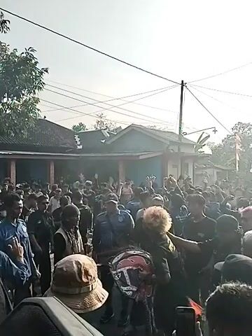A crowd of people is gathered outdoors in front of buildings. Many of the people are wearing dark clothing, and some appear to be associated with an organization or group, as indicated by matching uniforms or symbols. There are trees and power lines visible in the background, suggesting a semi-rural or suburban setting. The sun is shining, and the overall atmosphere appears to be one of a public event or gathering. One individual in the foreground has dyed blond hair and is being touched on the shoulder by someone else in the crowd. There is smoke or haze visible in the air near the center of the gathering. Some individuals are holding their hands up in the air, possibly in excitement or acknowledgement.