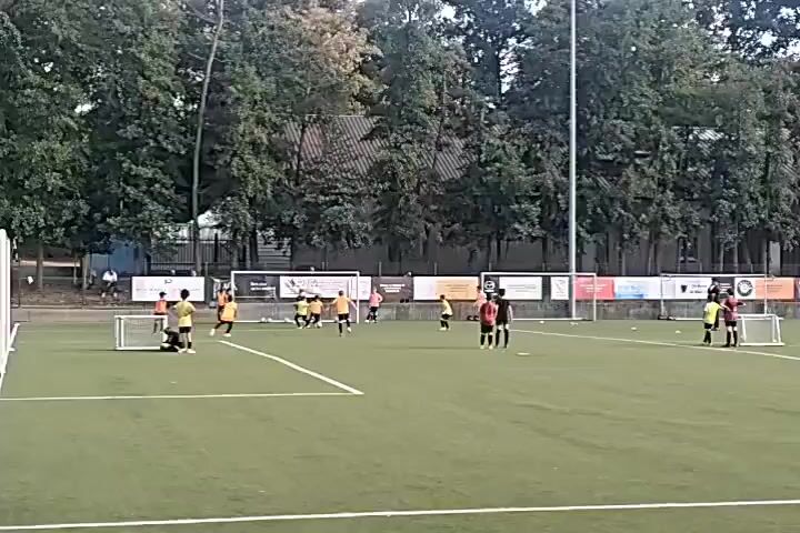 A group of young people in yellow and red jerseys are playing soccer on an artificial turf field. There are two small goals on the field. In the background, there are trees, a fence, and some buildings. The weather appears to be overcast.