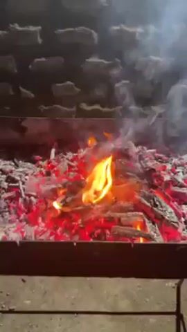 The video shows a close-up view of a charcoal fire in a grill. The embers are glowing red and there are flames rising from the pile of charcoal. Smoke is visible above the flames. The grill appears to be rectangular and made of metal. The background shows a textured, brick-like wall. There is ambient sound that may be music.
