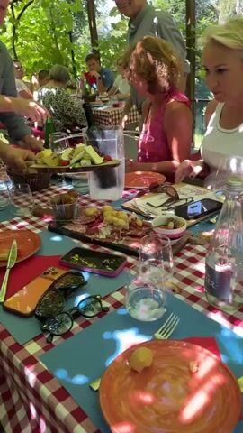 The video begins with a close-up shot of a table set for a meal. A red and white checkered tablecloth covers the surface, and several plates, glasses, and a bottle of green liquid are visible. In the foreground, a ceramic bowl filled with baked beans and pieces of meat is shown, with a spoon resting inside. The camera then pans to reveal slices of bread topped with some kind of spread and meat, arranged on a wooden board. A person's hand is visible holding a clipboard. The scene appears to be outdoors, under a canopy or roof structure, with dappled sunlight filtering through. The video then shifts to a selfie-style shot of a young man with dark hair wearing a white collared shirt. He is giving a thumbs-up to the camera, and a blurred background shows more of the outdoor dining area with a Swiss flag in the distance.