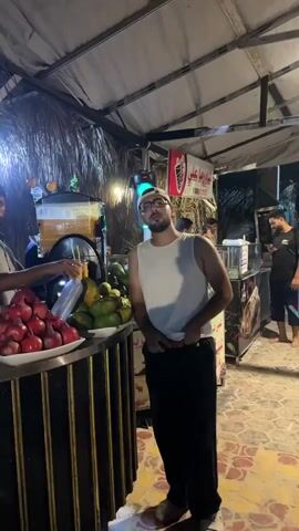 A man in a white tank top and black pants stands in front of a fruit stand. He is wearing glasses and a baseball cap. To his left, a fruit stand displays piles of pomegranates and papayas. Behind the fruit stand, several people are visible, some preparing drinks. The environment appears to be an outdoor market at night, with straw roofing and artificial lighting. The overall atmosphere is that of a bustling food stall.