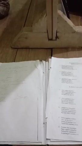 A group of people are sitting on the floor in a room with wooden floors and walls. Some people are reading books and writing in notebooks. One person is lying on their stomach with their head on the floor, appearing to be asleep. Another person is kneeling and looking at a book. The room appears to be a classroom or study area.