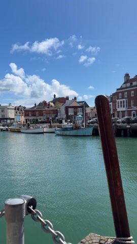 From a low angle, the camera pans from left to right along a harbor lined with buildings. The water is greenish-blue, reflecting the sunny sky with fluffy white clouds. On the left, the buildings are mostly red brick, with a couple of boats docked in front. As the camera pans right, the buildings become pastel shades of blue, white, pink, and yellow. Cars are parked along the pier in front of the buildings. A small yellow boat is docked further along. In the foreground, part of a pier structure is visible. A steel pole, rusty-colored, stands tall, and a chain is connected to the pier’s metal railing. The camera maintains a steady, low perspective, focusing on the colorful architecture and the calm harbor scene.