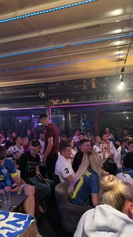 The video shows a gathering of people inside a dimly lit establishment, possibly a hookah lounge or bar. The ceiling is covered with blue and purple LED lights, and there are some decorative elements on the walls. The people are seated at tables, and many appear to be watching something, likely a screen not visible in the frame. Several individuals are holding mobile phones, and some are smoking from hookahs, evidenced by plumes of smoke. The overall atmosphere seems relaxed and social. People are dressed casually, with many wearing blue and yellow jerseys, suggesting a shared interest, possibly in a sports event. There are drinks and snacks on the tables. One person in the foreground is looking down at their phone. Another person, standing and wearing a maroon shirt, is looking towards the seated individuals. The lighting and the presence of hookahs suggest an indoor setting designed for entertainment and socializing.