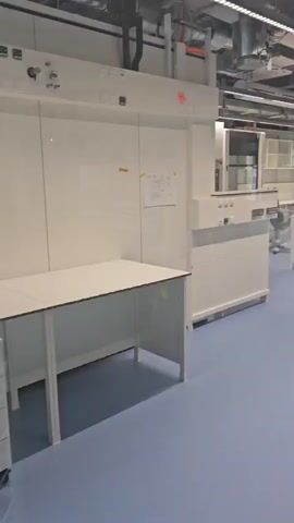 The video shows an empty, modern laboratory. The room is spacious with blue flooring and white lab benches and cabinets. Overhead, there are exposed pipes and ventilation systems, along with fluorescent lighting. There are no people or active experiments visible. A voice announces "Achtung. Hier spricht die Feuerwehr." which translates to "Attention. This is the fire department speaking."