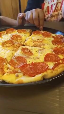 A close-up video shows two people, presumably a child and an adult, sitting at a wooden table and taking slices of a pepperoni pizza from a black metal tray. The pizza is round and cut into slices. It has a golden crust, a generous layer of melted cheese, and circles of pepperoni. Sprinkled herbs are visible on the pizza. One person is wearing a red shirt with white text and an image of palm trees, the other is wearing a black shirt with a design on it. The person who takes the slice is using their fingers to lift it off the tray. The person next to them is also reaching for a slice. The camera focuses closely on the pizza throughout the video. A wooden wall or panel is visible in the background.