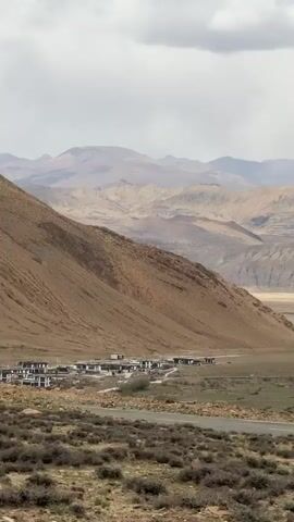 The video shows a landscape with a cluster of buildings nestled in a valley, surrounded by arid mountains. The buildings are small and have a uniform appearance, with light-colored walls and dark roofs.  The surrounding terrain is characterized by sparse vegetation, rocks, and dry dirt.  In the background, more mountains stretch out into the distance under a cloudy sky.  A road runs through the valley near the buildings.