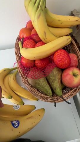 The video shows a fruit basket filled with bananas, clementines in red mesh bags, two pears, and two apples. A hand places another bunch of bananas from the side into the basket. The basket is made of woven material and is sitting on a white surface. The background is a plain white wall.