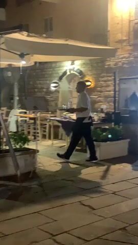 The video shows an outdoor dining area of a restaurant at night. Several tables are set with chairs and umbrellas overhead for shade. A man in a white shirt and black pants, presumably a waiter, walks across the scene carrying something. Two people are seated at a table in the background, facing away from the camera. Another person, also dressed in dark clothing, is visible in the far right background. Plants are in planters along the front of the dining area. The ground is paved with cobblestones.