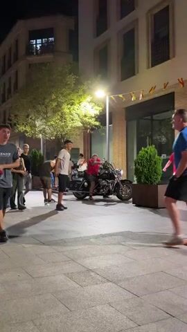The video shows an outdoor scene at night. In the foreground, a man with tattoos on his arm and wearing a red shirt and camouflage shorts is sitting on a motorcycle. He appears to be interacting with someone off-camera, possibly laughing or talking. To his left, another man in a white t-shirt is visible. Further in the background, other people are walking by, including two women and two men, one of whom is wearing a black t-shirt and denim shorts and walking away from the camera. The environment appears to be a street or sidewalk with brick buildings and foliage visible. The lighting suggests it is evening or night.
