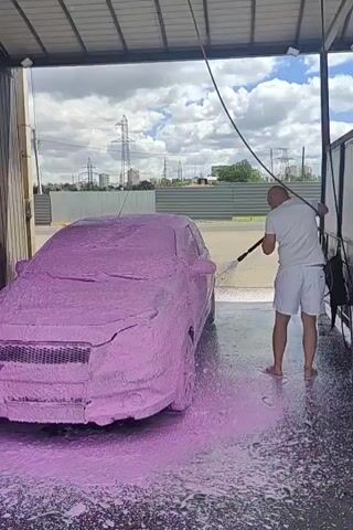 A man in white shorts and a white t-shirt is pressure washing a car covered in pink foam. The car is a sedan and is parked inside a covered car wash bay. The man is using a black pressure washer with a long wand. The foam is thick and covers the entire car. There is a lot of pink foam on the ground around the car. In the background, there are power lines and a fence. The sky is cloudy.