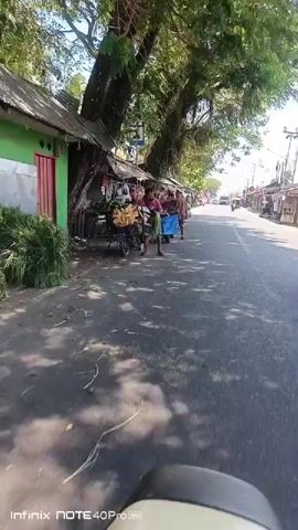 The video shows a street scene with several small shops or market stalls lining one side of the road. There are people present, some appearing to be vendors and others possibly customers or passersby. Several motorcycles are parked near the stalls. The stalls display various goods, including what appear to be fruits and vegetables. The road is paved and traffic, including motorcycles and a tricycle, can be seen in the distance. The scene is outdoors, under a sunny sky, with trees providing shade over some of the stalls. The camera appears to be mounted on a moving vehicle, likely a motorcycle, as indicated by the low angle and movement.