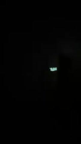 The video shows a dark room with several people. The faces and clothes of the people are not visible because of the darkness. Two screens are glowing with text on them, but the text is blurry and unreadable. One of the people is drinking from a cup or bottle.