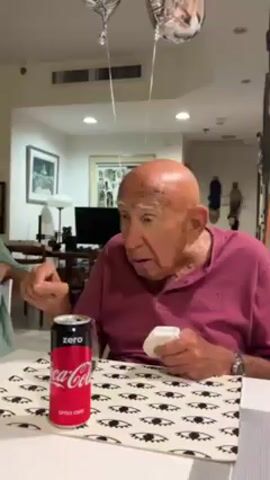 A video shows an elderly man sitting at a table with a can of Coca-Cola Zero next to him. He is wearing a pink polo shirt and has no hair. He is holding a small white item in his left hand and gesturing with his right. The table is covered with a white patterned tablecloth. Two silver balloons are visible in the background. The room appears to be a living or dining room with other furniture and decorations visible. The man seems to be speaking and reacting to something or someone off-camera.