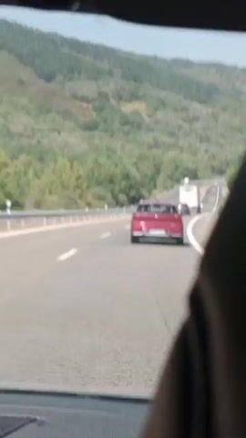 The video shows a view from inside a vehicle traveling on a highway. The highway is paved and has multiple lanes, marked by white lines. A red car is visible in the distance in the right lane, followed by a white truck in the left lane. The highway is surrounded by green, tree-covered hills. The sky is clear and blue. The video appears to be recorded during the daytime.