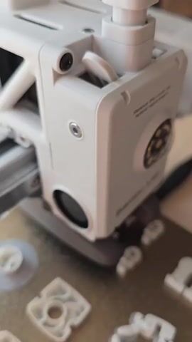 A 3D printer is shown printing white plastic parts on a textured build plate. The print head of the printer moves over the plate, extruding material to create the objects. Several small, intricately shaped parts are already printed and scattered across the build plate, some still attached to their support structures. A larger, more complex part is partially printed in the foreground. The background is blurred, but appears to be a wooden surface. The lighting suggests an indoor environment, possibly a workshop or desk.