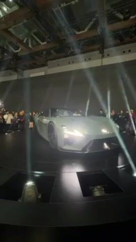 In a dimly lit indoor exhibition hall, a silver concept car is the focal point, rotating slowly on a black platform surrounded by spectators. Spotlights cut through the darkness, illuminating the sleek lines and sharp angles of the car's exterior, with the front headlights shining brightly. People are on both sides of the car with cameras and phones, capturing the car’s appearance. The exhibition hall is large and has a metal girder ceiling.