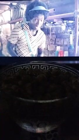 The video shows a hand reaching into a clear bowl of popcorn. In the background, a television screen displays a scene from a show or movie. The first frame shows a Black person with curly hair wearing a visor and a striped shirt. The second frame shows a light-skinned person with curly hair and bright eyes. The remaining frames show a person's hands interacting with an open book or papers on the television screen. The popcorn bowl is illuminated by the light from the television. The overall scene suggests someone is watching something on television and eating popcorn.