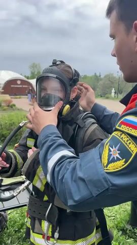 A young person wearing a full firefighter suit, complete with a gas mask, is being adjusted by an adult man in a Russian emergency services uniform. The man, whose uniform bears patches including the Russian flag and the emblem of the Ministry of Emergency Situations (МЧС РОССИИ), is carefully checking the fit of the gas mask. The person in the firefighter suit appears to be breathing into the mask, as indicated by the hose attached to it. In the background, a building and trees are visible under an overcast sky. The interaction suggests a training scenario or demonstration of firefighting equipment and procedures.