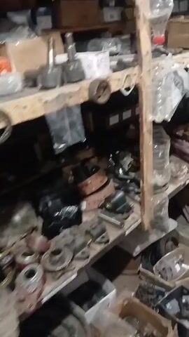 The video pans across a cluttered storage area filled with various auto parts and miscellaneous items. Shelves are packed with boxes, plastic bottles, and what appear to be engine components, filters, and metal pieces. Some items are hanging or propped against the shelves. The floor is also covered with more parts, including what looks like a circular grinding wheel. The lighting is dim, creating shadows throughout the space. The camera movement is shaky, suggesting it might be handheld.
