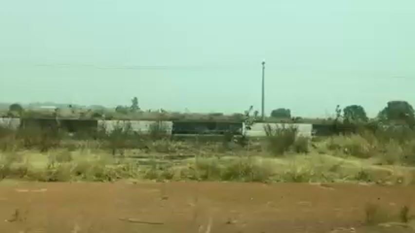 The video shows a view from a moving vehicle, likely a car or bus, looking out across a landscape of dry vegetation and scrubby trees. A road runs parallel to the camera's path, separated by a barrier painted with black and white stripes. Power lines stretch across the sky. The sky is a pale, hazy blue, suggesting a dusty or smoggy atmosphere. The foreground consists of reddish-brown soil and sparse, dry grass.  The overall impression is one of a dry, somewhat barren environment.