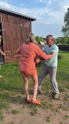 Two people are dancing outdoors in front of a wooden structure. The person on the left is wearing an orange outfit and orange Crocs, with their hands outstretched. The person on the right is wearing a light blue t-shirt, grey pants, and dark shoes, also with their hands outstretched in a dancing pose. The ground is covered in dirt and grass. A third person is partially visible in the background on the left, wearing a white t-shirt and ripped blue jeans. The sky is blue with some clouds.
