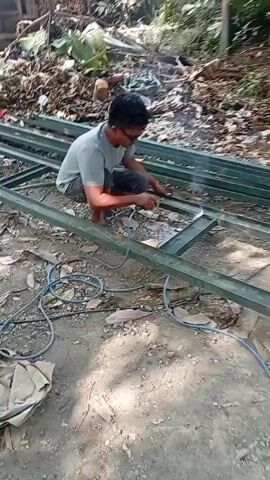 A person is welding metal beams in an outdoor setting. The person is kneeling and wearing glasses. There are sparks coming from the welding equipment. The ground is dirt and there are some leaves and debris. In the background, there are trees and foliage.