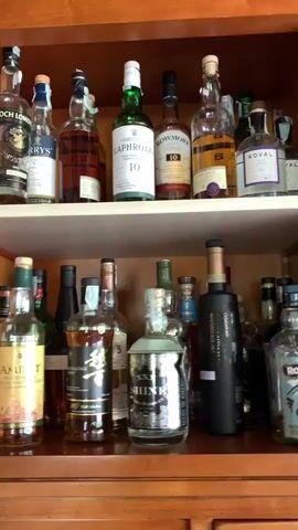 The media file shows a well-stocked liquor cabinet, likely located in a home, possibly in Castiglione d'Orcia, Italy. The cabinet is made of wood and has two shelves.

On the top shelf, there are various bottles of spirits, including what appears to be Scotch whisky, gin, and possibly tequila. Some of the visible labels include "Laphroaig 10," "Bowmore," and "Koval." The bottles are arranged in a somewhat orderly fashion, with some in the foreground and others further back.

The bottom shelf also displays a diverse collection of bottles. Prominently featured are bottles of gin, including "Hendrick's Gin." There are also bottles with labels such as "Caorunn," "Opthimus," and a yellow bottle with a stylized design. The arrangement here is also a mix of neat placement and some bottles positioned slightly behind others.

The lighting suggests it is daytime, though the specific time of day is not discernible. There are no people visible in the media file. The focus is entirely on the collection of alcoholic beverages. The wooden cabinet suggests a rustic or traditional aesthetic, which could align with a location in Italy. No specific weather conditions are visible as the scene is indoors. The overall impression is one of a curated collection of spirits, presented in a home setting.