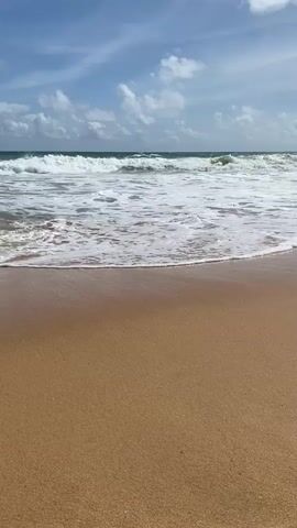 The video shows a beach with calm waves rolling onto the shore. The sand is a light brown color and the water is a clear blue. The sky is blue with a few white clouds. The waves are small and gentle, with white foam on top. The video is shot from a low angle, looking out at the ocean. There are no people or objects visible in the video. The overall scene is peaceful and serene.