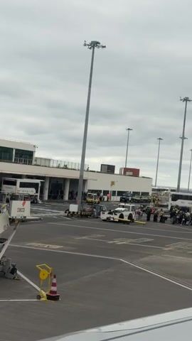 The video shows passengers disembarking from an aircraft at an airport.  A mobile stairway is positioned at the aircraft door. People with luggage are walking down the stairs and onto the tarmac. A bus labeled "My Way" is parked nearby, and passengers are boarding it. Airport infrastructure, including tall light poles and terminal buildings, is visible in the background. The sky is overcast.  The scene appears to be a typical airport operation during passenger arrival.