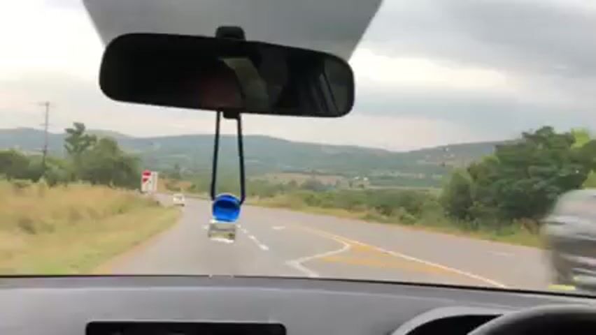 The video shows a drive along a road with a mountainous, rural landscape. The road is paved with yellow lines marking lanes. There's a car ahead. Green vegetation is visible on both sides of the road. The sky is cloudy and overcast.  The camera view is primarily from the dashboard looking out the windshield, and briefly shifts to the passenger side window. An air freshener hangs from the rearview mirror.