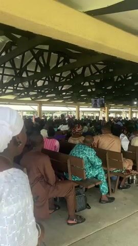 A wide shot shows a large gathering of people sitting on wooden benches under a covered outdoor structure. The structure has a yellow ceiling and a dark, crisscrossed roof support system. Many individuals are dressed in traditional African attire, with various colors and patterns visible, including browns, greens, purples, and pinks. In the background, several screens are mounted, displaying images of people, possibly a broadcast. A prominent yellow caution sign with black lettering is attached to one of the support beams. The ground appears to be concrete. The camera angle is slightly elevated, looking down at the seated crowd. Two men in traditional brown and black attire are walking towards the right side of the frame in the foreground. The overall atmosphere seems to be one of a public event or gathering.