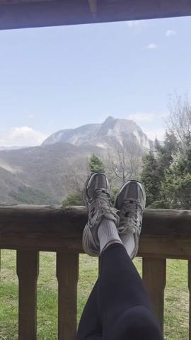 A person's legs and feet are prominently featured in the foreground of the image, propped up on a wooden railing, suggesting a relaxed posture on a porch or balcony. They are wearing grey and white New Balance athletic shoes with white socks and black leggings or pants; the "N" logo is visible on the side of the shoes.

Beyond the railing, a vast mountainous landscape unfolds under a clear, bright sky with scattered clouds. In the midground, various trees are visible; some deciduous trees appear bare, indicating late winter or early spring, while evergreen trees add patches of deep green. A large, rocky mountain dominates the background, showcasing rugged terrain and sparse vegetation on its slopes. The overall impression is one of peaceful relaxation amidst a scenic natural environment, likely in a mountainous region such as Laki, Bulgaria, during daytime.