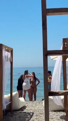 A vertical video on a bright, clear day, shows four women standing on a pebble beach at the edge of the blue sea. The women are talking and laughing. Two canopies with white curtains are visible, framing the scene. Two of the women are in swimwear, one in a white top and shorts, and the other in a black top and shorts. The distant horizon shows calm water and a clear sky. A wooden structure is on the right, also framing the scene.