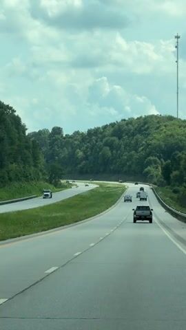 The video shows a highway with multiple cars driving on it. The highway is surrounded by lush green trees on both sides. The sky is partly cloudy with a blueish tint. A tall cell tower is visible on the right side of the screen. The camera appears to be mounted inside a moving vehicle, likely capturing the view from the passenger seat or dashboard. The overall atmosphere is serene and suggests a scenic drive through a rural or mountainous area.