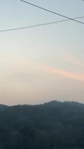 The video shows a blurry, low-angle shot of a dark utility pole in the foreground, angled to the right. The pole is partially obscured by motion blur and has a yellow stripe near the bottom. Above the pole, two thin wires run horizontally across the frame. In the background, a line of dark, tree-covered hills stretches across the horizon. The sky above the hills is a pale gradient of blue and pink, suggesting either sunrise or sunset. The camera appears to be moving, causing the motion blur. There are no people or distinct actions visible in the frame.