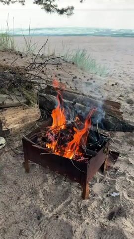 A campfire burns in a metal brazier on a sandy beach. The fire is composed of logs and branches, with orange flames and white smoke rising into the air. In the background, the ocean is visible with gentle waves and a sandy shore. There are also some pine needles and branches scattered around the brazier. The overall scene depicts a peaceful outdoor setting.