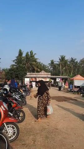 A woman in a black hijab and a floral dress walks across a dusty, open market area. Several motorcycles are parked on the left side of the frame. In the background, there are market stalls with umbrellas, and people are browsing and interacting. Palm trees and a bright blue sky are visible above. The scene appears to be an outdoor market on a clear day.