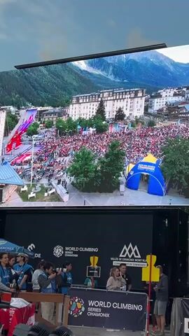 The video captures an outdoor event with a large crowd gathered in a plaza. A massive screen displays an aerial view of the same plaza, showing buildings, a mountain backdrop, and many people. In the foreground, a stage is set up with a banner that reads "WORLD CLIMBING SERIES CHAMONIX." Several people are standing on the stage, some appearing to be organizers or commentators, while others are holding cameras. The crowd is watching the screen, suggesting a live broadcast or replay of a climbing event. The overall atmosphere seems to be one of excitement and anticipation.