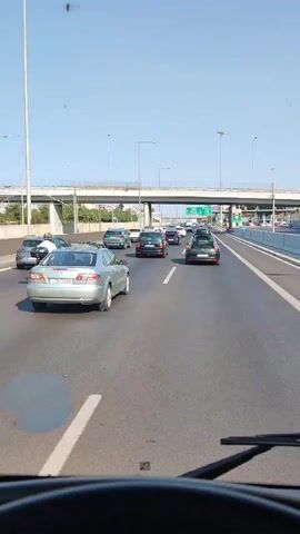 This is a point-of-view video from a vehicle driving on a multi-lane highway. The scene is bathed in bright daylight, suggesting a clear and sunny day. The road is populated with several cars, indicating moderate traffic. The cars are predominantly silver and black, and they are moving in the same direction.

In the foreground, the dashboard and windshield wipers of the vehicle are visible, framing the view of the road ahead. A light silver sedan is prominently featured in the left lane, with its license plate clearly visible: "IZB 7288". Other cars are spread across the lanes, with some further in the distance.

Overhead, a large concrete overpass crosses the highway, with structural supports visible on the side. Streetlights line the highway, suggesting it's a well-maintained road. Trees and vegetation can be seen on the embankment beyond the overpass, adding a touch of nature to the urban infrastructure. The sky is a clear, vibrant blue, with no clouds in sight. The overall impression is one of a typical daytime commute or drive through a developed area, likely a city or its outskirts, given the presence of the highway and overpass. The video appears to be recorded in Greece, based on the context provided. No specific emotional tone is conveyed, beyond the normalcy of highway travel. There are no other people visible other than those potentially inside the vehicles.