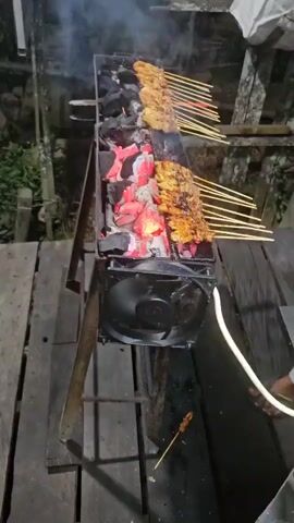 A row of satay skewers is being grilled over hot coals. The skewers are arranged in two parallel lines on a metal grill. Below the grill, a fan is attached to a metal frame, presumably to stoke the coals. The coals are glowing red and orange, with smoke rising from them. The scene appears to be outdoors, possibly at night, given the lighting. There are wooden planks visible beneath the grill. The satay appears to be chicken or a similar type of meat, with a golden-brown color from the grilling. A white cord is visible to the right of the fan.