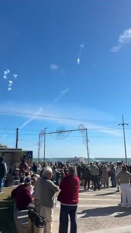 The video captures an outdoor daytime scene with a large gathering of people along a waterfront. Under a clear blue sky, numerous white smoke puffs and explosions are visible, indicating a daytime fireworks or pyrotechnic display. The crowd, mostly adults, stands and sits, observing the sky.

In the foreground, a decorative archway featuring wireframe seahorse figures and a central shell-like design spans the path leading towards the water. Power lines crisscross the upper sky. In the background, beyond the crowd, a body of water (likely the ocean) is visible, and a white truck with 'DOURO' written on it can be seen. A white road sign with a red triangle is present on the left. The bright sunlight and the gathering suggest a public event in a coastal setting, consistent with Vila Nova de Gaia, Portugal.