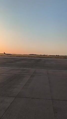 The media file captures a wide shot of an airfield during sunset. The sky transitions from a pale orange near the horizon to a clear blue overhead. In the distance, an airplane is visible on the tarmac, appearing to be stationary or moving slowly. Beyond the runway, a blurred line of buildings suggests the presence of an airport or adjacent urban area. The foreground is dominated by a concrete runway or taxiway, marked with white lines. The lighting indicates it is late afternoon or early evening, with the low sun casting a warm glow. The scene appears calm and still, with no visible people or other vehicles in the immediate vicinity. The overall impression is one of quiet solitude at a transitional time of day.