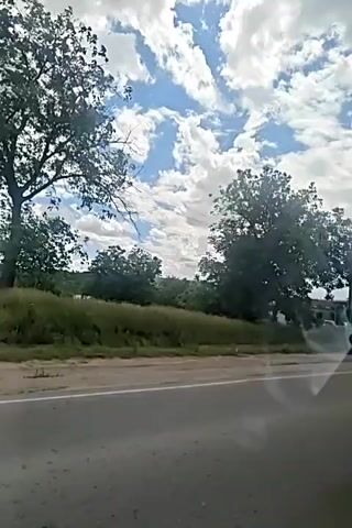 The video captures a drive-by perspective of a rural or suburban roadside scene. The sky is filled with dramatic, white, puffy clouds against a bright blue backdrop. In the foreground, a paved road is visible with a white line marking the lane.

To the left of the road, there's a grassy embankment with tall, green grass and some vegetation. A large, leafy tree dominates the middle ground. Behind the tree, several structures are visible, including a white building with a grey roof and some dark, box-like structures, possibly garages or storage units. Power lines and utility poles are prominent throughout the scene, with one particularly large pole to the right.

Several vehicles are parked or driving on the side of the road. A red SUV is parked prominently. Further down, a white sedan is visible, and what appears to be a black SUV or truck is parked. In the distance, more buildings and vehicles can be seen, suggesting a small commercial or industrial area. The overall impression is a sunny day with scattered clouds, showing a typical roadside environment with some parked vehicles and infrastructure.