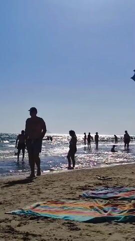 The video shows a crowded beach on a sunny day. The camera is positioned on the sand, facing the ocean. In the foreground, there are two towels laid out on the sand. In the middle ground, people are walking along the shore and wading in the shallow water. The water is clear and reflects the bright sun. In the background, more people are visible in the water and on the beach, with a hazy coastline visible in the far distance. The sky is a clear, bright blue. The overall atmosphere is one of a typical summer day at the beach, with people enjoying the sun and the sea.