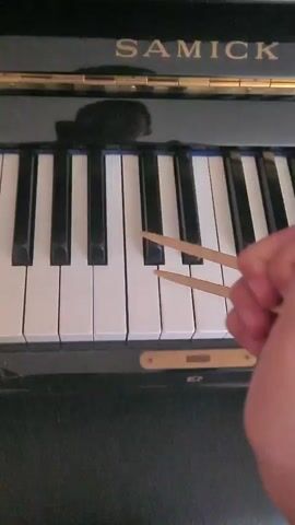 A person's hand holds two chopsticks over the black and white keys of a piano. The piano has the brand name SAMICK visible at the top. The chopsticks are positioned to strike the keys, and the video shows them playing a short melody by hitting the white keys. The background is dark and appears to be the interior of the piano.
