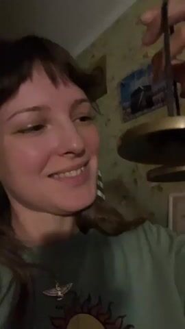 The video shows a close-up of a young woman's face as she smiles and looks slightly to her right. She is wearing a light green t-shirt with a sun design on it and a pendant. In her right hand, she holds a small, brass bell with a string attached. The bell is being gently shaken, creating a faint ringing sound. The background is out of focus, but appears to be an indoor setting with patterned wallpaper and a framed picture on the wall. The lighting is dim.