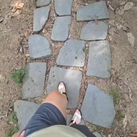 The video begins with a close-up aerial view of a person's feet and legs as they walk along a path made of irregular flat stones. The path is surrounded by dirt, leaves, and some sparse green vegetation. The person is wearing dark blue shorts and white sandals with black straps. As the video progresses, the camera angle shifts slightly, giving a more dynamic perspective of the walk. The person's stride is casual, and they appear to be moving at a moderate pace. The lighting suggests an outdoor environment, possibly a park or trail, with sunlight filtering through trees, creating dappled shadows on the ground. The video concludes as the person continues to walk further down the path, leaving the initial stone section behind and transitioning onto a dirt and gravel surface.